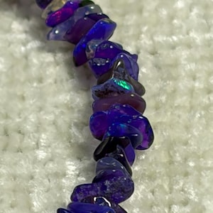 May include: A close-up of a bracelet made of irregular, polished, dark purple gemstones with iridescent flashes of color. The gemstones are strung together, creating a unique and eye-catching piece of jewelry. The background is a soft, white, textured surface.