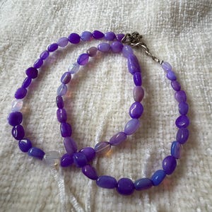 Könnte beinhalten: Eine Halskette mit ovalen Perlen in verschiedenen Lilatönen, von tiefem Violett bis hell Lavendel. Die Perlen sind aufgefädelt und bilden ein doppelreihiges Design. Die Halskette wird auf einer weißen, strukturierten Oberfläche gezeigt.
