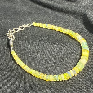 Può includere: Un delicato braccialetto con piccole perline rotonde giallo-verdi infilate su una catena e chiusura color argento. Le perline hanno una sottile iridescenza. Il braccialetto è esposto su una superficie scura e strutturata.