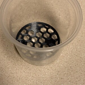 May include: A clear plastic container with a black, honeycomb-patterned insert at the bottom. The container is cylindrical and sits on a speckled, light-coloured surface. The insert appears to be made of plastic.