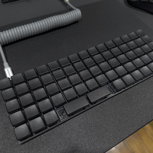 May include: A black mechanical keyboard with square keycaps and a coiled gray cable. The keyboard is on a dark surface, with a mouse and other items in the background.