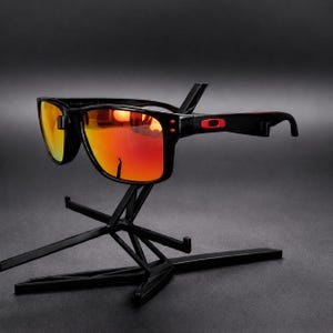 Pode incluir: Óculos de sol pretos com armação retangular e lentes refletoras em tons vibrantes de laranja e vermelho. As hastes exibem um logótipo retangular vermelho proeminente perto das dobradiças, criando um contraste marcante com a armação.