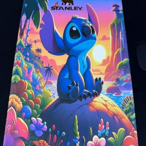 Op de afbeelding: Een rechthoekige productdoos met een levendige, kleurrijke illustratie van het blauwe personage Stitch zittend op een rots. De scène bevat een tropisch landschap met palmbomen, bloemen en een zonsondergang. Het Stanley-logo staat bovenaan gedrukt.