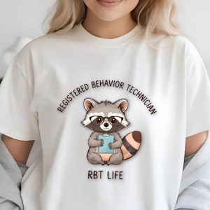 Może przedstawiać: Biały t-shirt z krótkim rękawem z grafiką przedstawiającą szopa pracza w okularach z czarnymi oprawkami, trzymającego jasnoniebieską podkładkę z klipsem. Tekst powyżej brzmi REGISTERED BEHAVIOR TECHNICIAN, a poniżej RBT LIFE.