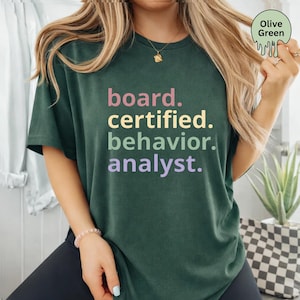 Pode incluir: Uma t-shirt de manga curta verde-oliva com o texto "board. certified. behavior. analyst." impresso em quatro cores distintas: rosa, amarelo, verde-claro e roxo-claro, disposto em quatro linhas na parte frontal da peça de vestuário.