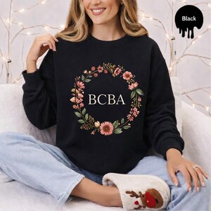 Könnte beinhalten: Ein schwarzes Langarm-Sweatshirt mit einem kreisförmigen Blumenkranz-Motiv in Rosa-, Orange- und Grüntönen. Die Buchstaben BCBA sind in Weiß in der Mitte des Kranzes aufgedruckt. Das Sweatshirt hat einen Rundhalsausschnitt und gerippte Bündchen.