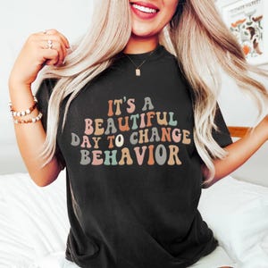 Op de afbeelding: Zwart T-shirt met de tekst "It's a Beautiful Day to Change Behavior" in kleurrijke, retro letters. Het shirt heeft een ronde hals en korte mouwen. De tekst is in tinten roze, oranje, blauw en groen.