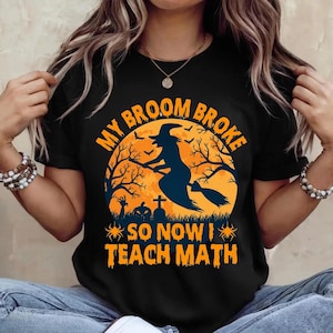 May include: A black t-shirt featuring a graphic of a witch flying on a broomstick against a large orange moon. The text reads "MY BROOM BROKE SO NOW I TEACH MATH" in orange letters. The design includes silhouettes of trees, a pumpkin, and a tombstone.