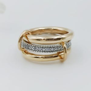 Op de afbeelding: Een goudkleurige ring met een drievoudig bandontwerp. De middelste band is bezet met kleine, heldere edelstenen. De ring heeft een uniek, verweven ontwerp met een gepolijste afwerking, geschikt voor dagelijks gebruik of speciale gelegenheden.