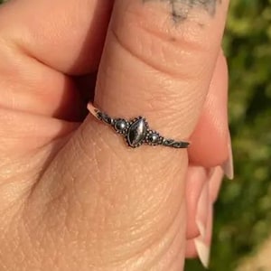 May include: A silver ring with an oval, dark-colored stone at the center. The ring has a delicate, ornate design with small, round accents on either side of the central stone. The ring is worn on a finger.