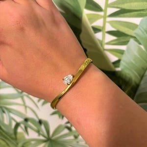 Puede incluir: Una pulsera dorada con un diamante en forma de pera. La pulsera tiene un diseño curvo y se lleva en la muñeca. El fondo presenta hojas verdes.