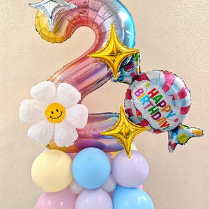 May include: A colorful balloon arrangement for a birthday celebration. The centerpiece is a large number 2 balloon with a rainbow gradient. It is decorated with star-shaped balloons, a daisy with a smiley face, and a balloon that says "HAPPY BIRTHDAY!"