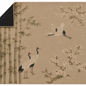 May include: A rectangular mat with a tan background featuring a printed scene of two standing cranes, one flying crane, bamboo stalks, and flowering branches. The top left corner is folded over to reveal a solid black underside with a thin tan border.
