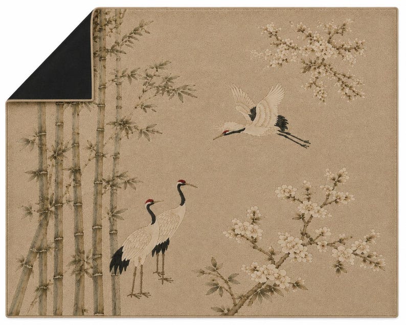 May include: A rectangular mat with a tan background featuring a printed scene of two standing cranes, one flying crane, bamboo stalks, and flowering branches. The top left corner is folded over to reveal a solid black underside with a thin tan border.