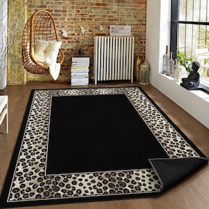 Pode incluir: Um tapete retangular com um painel central preto s&oacute;lido rodeado por uma borda larga com um padr&atilde;o de estampa de leopardo em tons de bege e castanho. O tapete tem um verso cinzento escuro vis&iacute;vel no canto onde a borda est&aacute; dobrada.