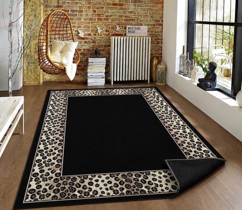 Pode incluir: Um tapete retangular com um painel central preto s&oacute;lido rodeado por uma borda larga com um padr&atilde;o de estampa de leopardo em tons de bege e castanho. O tapete tem um verso cinzento escuro vis&iacute;vel no canto onde a borda est&aacute; dobrada.