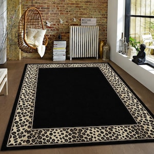 Pode incluir: Um tapete retangular com um painel central preto s&oacute;lido, emoldurado por uma borda larga com um padr&atilde;o de leopardo em tons de bege e castanho escuro. O tapete tem um contorno preto fino e est&aacute; colocado sobre um piso de madeira clara e lisa.
