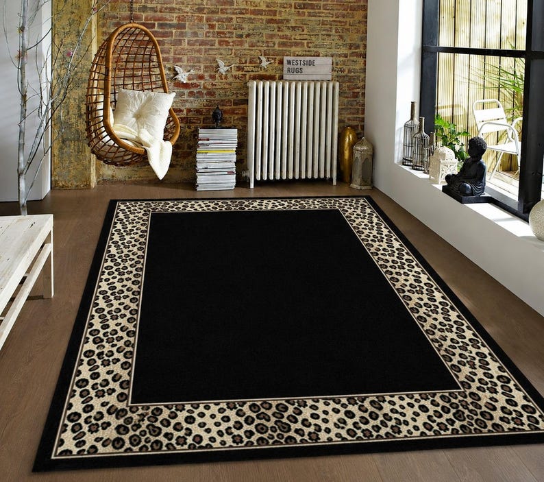 Pode incluir: Um tapete retangular com um painel central preto s&oacute;lido, emoldurado por uma borda larga com um padr&atilde;o de leopardo em tons de bege e castanho escuro. O tapete tem um contorno preto fino e est&aacute; colocado sobre um piso de madeira clara e lisa.