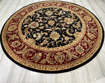 Pode incluir: Tapete redondo com um centro preto com padrões florais dourados e bege. Uma borda larga em bordô e dourado circunda o centro, com uma borda externa preta. O tapete é projetado para uso interno.