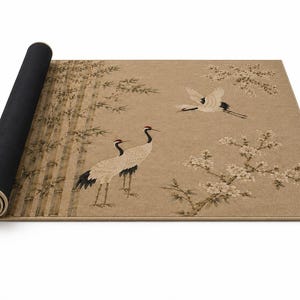 May include: A rectangular area rug with a tan background featuring a printed design of two standing cranes, one flying crane, bamboo stalks, and flowering branches. The rug is partially rolled up, revealing a solid black underside and a tan edge binding.