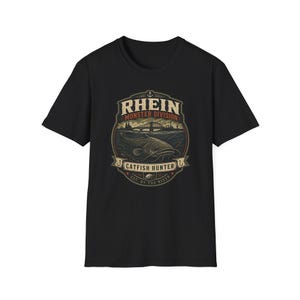 Peut inclure: Un t-shirt noir à manches courtes avec une impression graphique représentant un poisson-chat dans l'eau sous un pont. Le texte indique RHEIN MONSTER DIVISION, CATFISH HUNTER et EST. BY THE RIVER. Le motif inclut une ancre et deux hameçons.