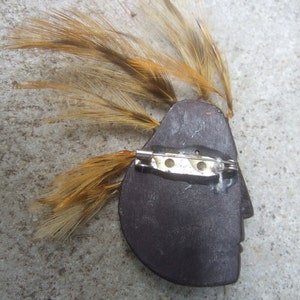 May include: A wooden brooch with a profile of a face and a brown feather headdress. The brooch has a silver pin and catch.