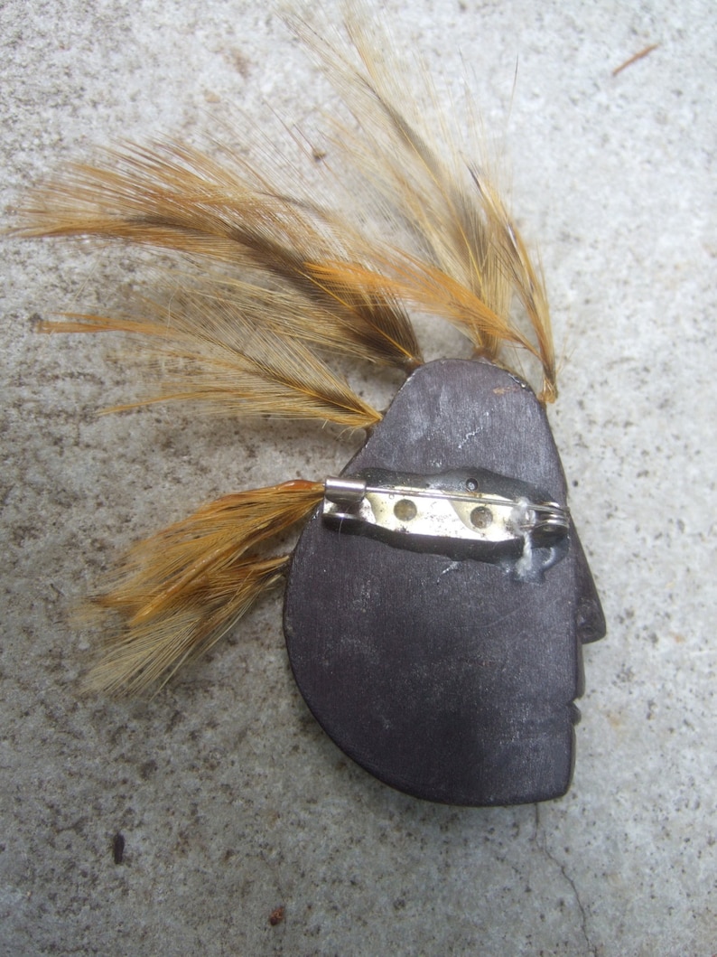 May include: A wooden brooch with a profile of a face and a brown feather headdress. The brooch has a silver pin and catch.
