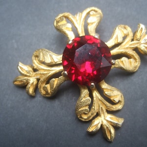 May include: A gold-tone brooch with a large, red, faceted gemstone in the center. The brooch is shaped like a stylized flower with four petals.