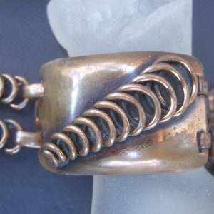 Pode incluir: Um bracelete de cobre com um design decorativo de círculos entrelaçados. O bracelete é feito de duas seções que se conectam com um fecho.