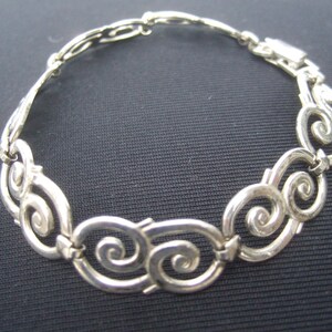 May include: A silver bracelet with a swirling, ornate design. The bracelet is made of linked, interconnected loops.