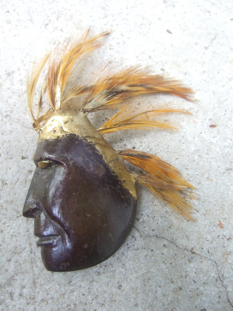 May include: A brown wooden mask with a gold painted design and a feather headdress. The mask has a stylized human face with a long nose and closed eyes.