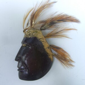 May include: A wooden mask with a profile view of a face, painted brown and gold. The mask has a headdress of brown feathers.