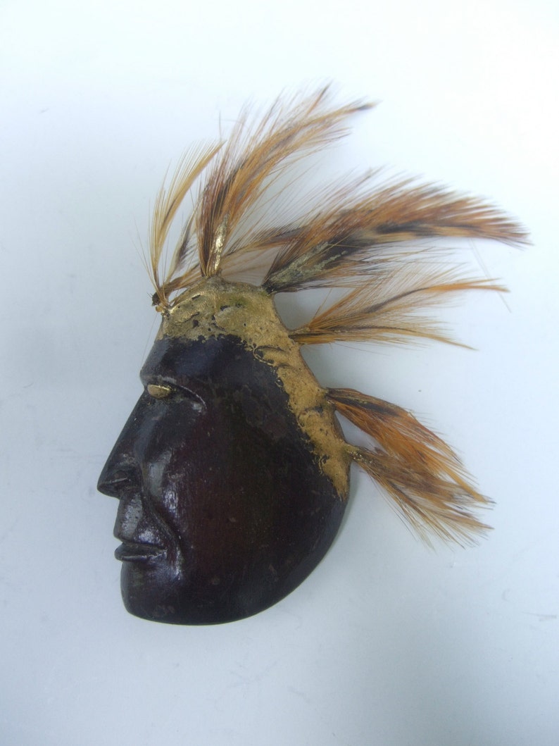 May include: A wooden mask with a profile view of a face, painted brown and gold. The mask has a headdress of brown feathers.