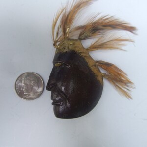 May include: A wooden Native American mask with a gold painted headdress and brown feathers. The mask has a carved face with a serious expression.