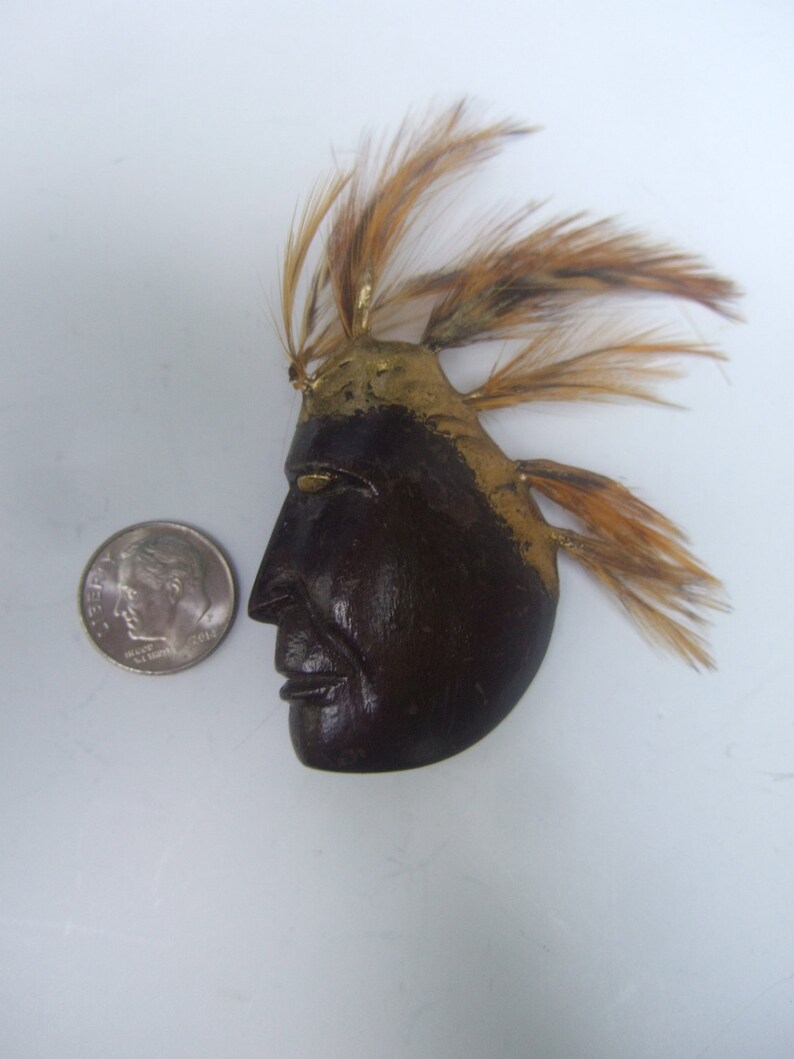 May include: A wooden Native American mask with a gold painted headdress and brown feathers. The mask has a carved face with a serious expression.