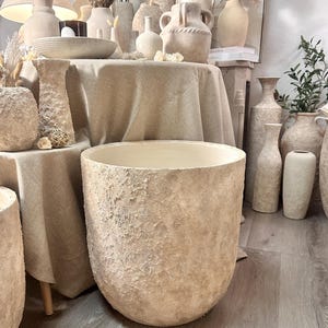 May include: A large, cylindrical floor planter with a rough, textured, beige stone-like finish. The exterior surface features a mottled, uneven appearance with light and dark cream tones, while the interior is smooth and off-white.