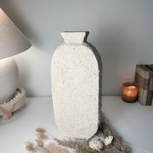 May include: A tall, off-white vase with a flat, oval-shaped body and a narrow neck. The surface of the vase features a heavy, rough, and textured finish that covers the entire exterior, creating a tactile appearance with visible ridges and bumps.