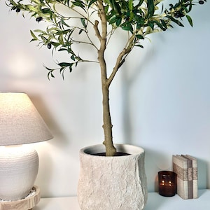 May include: An artificial olive tree with green and variegated leaves and small dark olives, planted in a textured, off-white, cylindrical stone-like planter. The tree has a central brown trunk that branches out into a full canopy of foliage.