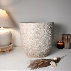 May include: A large, cylindrical planter with a rough, textured surface in shades of beige and off-white. The exterior features a thick, uneven application of material, creating a tactile, weathered appearance with a wide, open top and a rounded base.