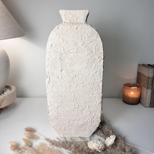 May include: A tall, off-white vase with a flat, oval-shaped body and a narrow neck. The surface of the vase features a thick, textured, stucco-like finish with visible brush strokes and uneven patterns throughout the entire exterior of the object.