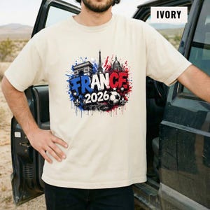 May include: Ivory-colored t-shirt featuring a graphic design with the word "FRANCE" in blue, and "2026" with a soccer ball. The design also includes the Eiffel Tower, Arc de Triomphe, and other architectural elements in red and blue.