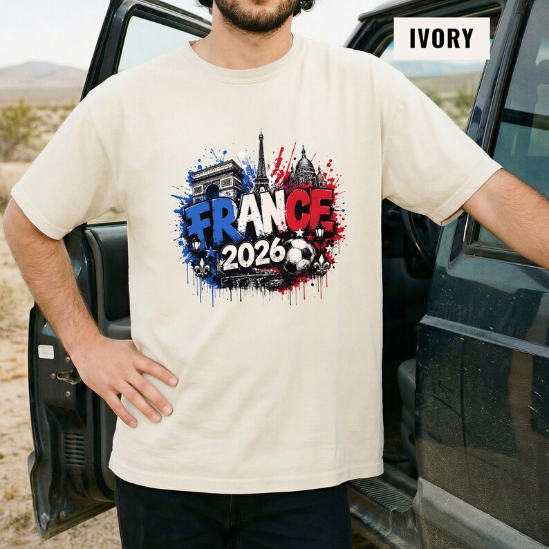 May include: Ivory-colored t-shirt featuring a graphic design with the word "FRANCE" in blue, and "2026" with a soccer ball. The design also includes the Eiffel Tower, Arc de Triomphe, and other architectural elements in red and blue.