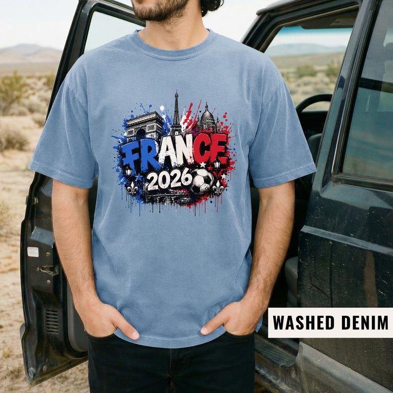 May include: A light blue t-shirt with a graphic design featuring the Eiffel Tower, Arc de Triomphe, and a soccer ball. The word "FRANCE" is displayed in red, white, and blue, with "2026" below. The shirt is labeled "WASHED DENIM".
