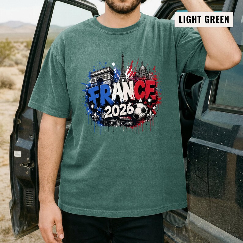 May include: Light green t-shirt with a graphic design featuring the word "FRANCE" in blue and red, the year "2026", and images of the Eiffel Tower, Arc de Triomphe, and a soccer ball. The shirt is a short-sleeved crew neck.
