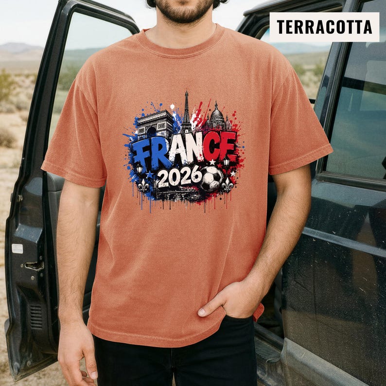 May include: Terracotta-colored t-shirt featuring a graphic design with the word "FRANCE" in blue, white, and red, along with the year "2026" and a soccer ball. The design includes illustrations of the Eiffel Tower, Arc de Triomphe, and other architectural elements.