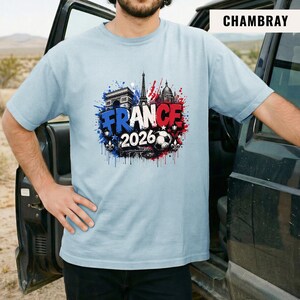 May include: Light blue t-shirt with a graphic design featuring the word "FRANCE" in blue and red, the year "2026", and iconic French landmarks like the Eiffel Tower and Arc de Triomphe. A soccer ball is also included in the design.