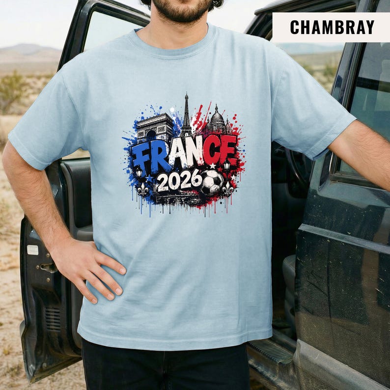 May include: Light blue t-shirt with a graphic design featuring the word "FRANCE" in blue and red, the year "2026", and iconic French landmarks like the Eiffel Tower and Arc de Triomphe. A soccer ball is also included in the design.