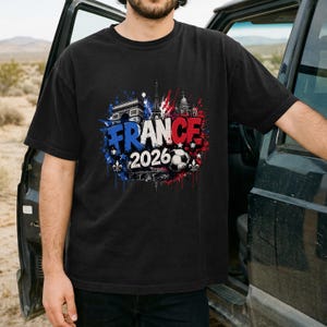 May include: Black t-shirt featuring a colorful graphic design with the text "FRANCE 2026". The design incorporates elements of the French flag, a soccer ball, and iconic landmarks like the Eiffel Tower and Arc de Triomphe.