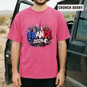 May include: A short-sleeved, pink t-shirt featuring a graphic design with the word "FRANCE" in blue and red, the year "2026", and images of the Eiffel Tower, Arc de Triomphe, and a soccer ball. The shirt is a solid color.