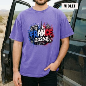 May include: Violet short-sleeve t-shirt featuring a graphic design with the text "FRANCE 2026" in white, blue, and red, along with images of the Eiffel Tower, Arc de Triomphe, and a soccer ball. The shirt is a solid violet color.
